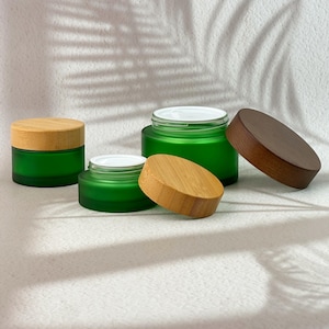 May include: Three round cosmetic jars made of frosted green glass. Two jars have light-colored bamboo lids, one of which is placed beside its jar. The third jar features a dark brown wooden lid and is shown with its lid resting separately.
