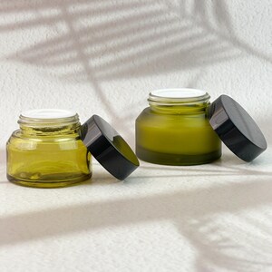Puede incluir: Dos pequeños frascos de vidrio verde oliva con tapas negras, una tapa está abierta. Los frascos tienen un interior blanco y están llenos de una sustancia verde claro. Los frascos están sobre una superficie blanca con una sombra de hojas en el fondo.