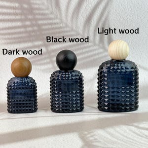 May include: Three dark blue glass bottles with textured surfaces and wooden ball stoppers. The stoppers are labeled "Dark wood", "Black wood", and "Light wood". The bottles are arranged from smallest to largest.