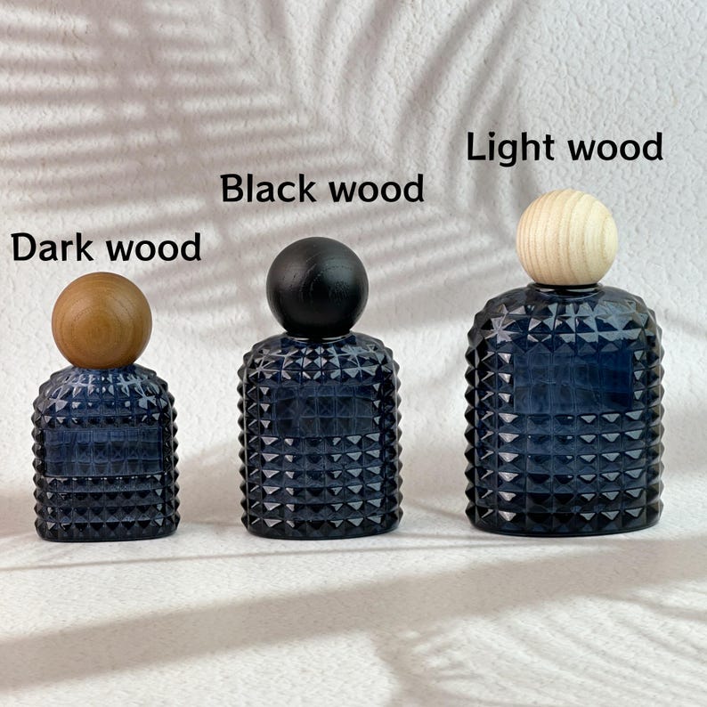 May include: Three dark blue glass bottles with textured surfaces and wooden ball stoppers. The stoppers are labeled "Dark wood", "Black wood", and "Light wood". The bottles are arranged from smallest to largest.