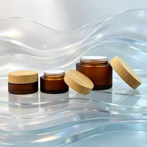 May include: Three amber-colored glass jars with light-colored wooden lids. Two jars are closed with lids, while one larger jar is open with its lid resting beside it. The jars have a smooth, matte finish and a cylindrical shape.
