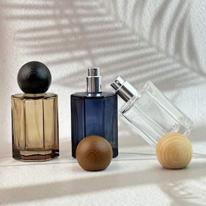 May include: Three glass perfume bottles in amber, blue, and clear. Each has a silver atomizer and a wooden cap or sphere. The amber bottle has a dark wooden cap. The blue bottle has a wooden sphere. The clear bottle is leaning.