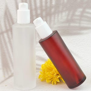 May include: Two cylindrical bottles with white pump tops. One bottle is frosted, the other is a deep red. A yellow flower is in the foreground. The bottles are likely for skincare or cosmetic products.