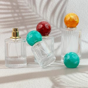 May include: Clear glass perfume bottles with gold-colored sprayers and decorative, spherical stoppers in red, teal, and orange. The bottles have a ribbed design and are arranged on a white surface.