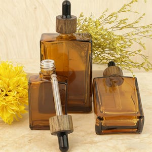 May include: Amber glass dropper bottles with wooden caps. The bottles are square-shaped and come in various sizes. One bottle has a dropper inserted, while others have the wooden cap on. The background includes yellow flowers and dried plants.