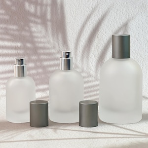 May include: Three frosted glass perfume bottles of varying sizes with silver spray nozzles and dark gray cylindrical caps. The bottles have a rounded, bulbous shape and a smooth, translucent finish, arranged in a row on a textured surface.