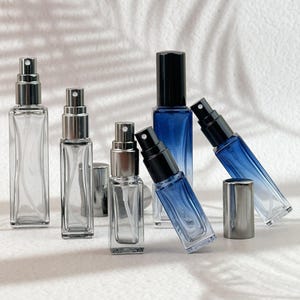 Puede incluir: Un surtido de botellas de spray rectangulares de vidrio con pulverizadores plateados y negros. Algunas botellas son transparentes, mientras que otras tienen un color azul degradado. Las botellas están dispuestas sobre una superficie blanca.