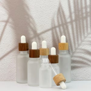May include: A collection of frosted glass dropper bottles with wooden and white tops. The bottles are arranged on a white surface, with a shadow of a plant in the background. One bottle has its dropper exposed.
