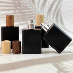 Pode incluir: Várias garrafas de perfume retangulares pretas com tampas de madeira e borrifadores prateados são exibidas. Algumas garrafas têm tampas de madeira, outras mecanismos de borrifador prateados. As garrafas estão dispostas em uma superfície branca.