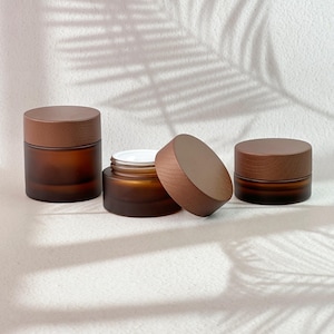 May include: Three cylindrical cosmetic jars made of dark amber frosted glass, each topped with a smooth, circular wooden lid. One jar is shown open with a white inner lid visible, while the other two jars remain closed with their wooden lids secured.
