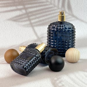 Four navy blue diamond cut glass spray bottles, two tilted to show spray pumps, with dark/black/light wood caps on white textured background.