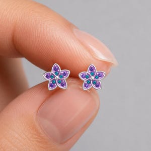 May include: A pair of silver stud earrings shaped like five-petaled flowers. Each petal is filled with purple enamel and accented with small, embedded teal-colored stones. The center of each flower features a small, raised silver dot detail.