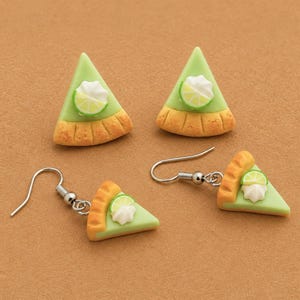 May include: Two pairs of lime pie slice earrings. One pair features stud posts, while the other pair includes silver-toned metal fishhook wires. Each triangular piece has a light green filling, a tan crust edge, and a white dollop of cream.