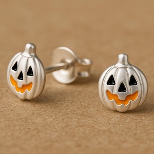 May include: A pair of silver stud earrings shaped like jack-o'-lantern pumpkins. Each earring features a metallic silver finish with black triangular eyes and a nose, complemented by a bright orange enamel mouth with a jagged tooth design.