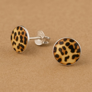 May include: A pair of round stud earrings featuring a tan and black leopard print pattern. Each earring is set in a silver-toned metal frame and includes a matching silver-toned post and butterfly back closure for secure attachment to the earlobe.