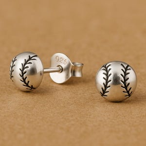 May include: A pair of silver stud earrings shaped like baseballs, featuring engraved black stitching details. The earrings have a post back closure and are stamped with the number 925, indicating sterling silver composition for these small accessories.
