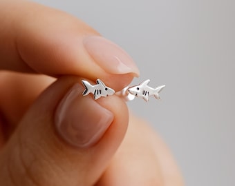 May include: A pair of small, polished silver stud earrings shaped like sharks. Each earring features a minimalist shark silhouette with a tiny engraved eye and gill details, designed with a post back for secure attachment to the earlobe.