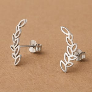 May include: A pair of silver-colored leaf-shaped stud earrings. Each earring features a curved design of connected leaf shapes. The earrings are displayed on a light brown surface.