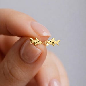 May include: A pair of small gold stud earrings shaped like sharks. Each earring features a polished gold finish with a simple cutout design for the eye and gill slits, showcasing a minimalist silhouette of a shark facing toward the center.