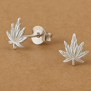 May include: A pair of silver-colored stud earrings. Each earring is shaped like a cannabis leaf with detailed veining. The earring posts are visible, and the background is a solid tan color.