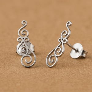 May include: A pair of silver-colored stud earrings. Each earring features an openwork design with swirling, scroll-like patterns. The earrings are displayed on a neutral, tan-colored surface.