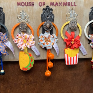 May include: Five door hangers with different designs. Each hanger has a ribbon with a bow, a charm, and a bell. The charms are a taco, a french fry container, a flower, a butterfly, and a pretzel. The ribbon on each hanger says "House of Maxwell".