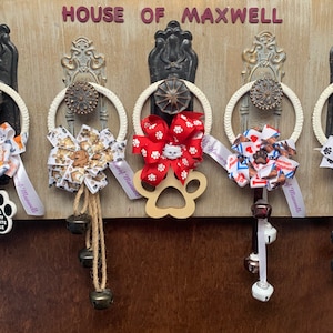May include: Five door hangers with paw print designs. Each hanger has a white rope loop, a bow with a dog-themed print, and a wooden paw print charm. The bows are red, white and blue, red and white, brown and white, and black and white. The sign above the hangers reads "House of Maxwell".