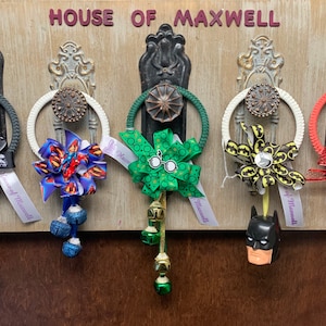 May include: Five door hangers with different themed bows and charms. The bows are green, blue, yellow, red, and black. The charms are bells, a Batman figure, and a red superhero figure. The door hangers are labeled "House of Maxwell".