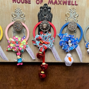 May include: Five colorful door hangers with different Disney character themes. Each hanger features a bow, a character charm, and a set of bells. The hangers are labeled "House of Maxwell".