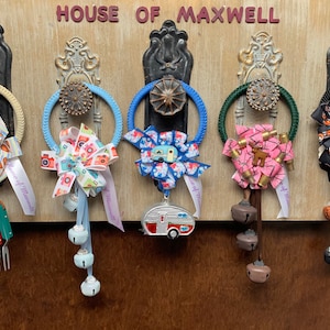 May include: Five door wreaths with different colored ribbons and bells hanging from a wooden door with the text "House of Maxwell" above them.