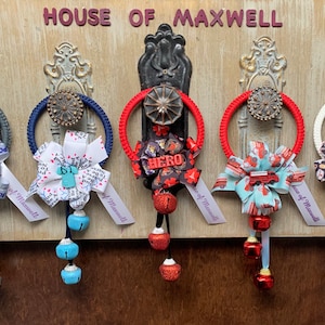 May include: Five door wreaths with different themed ribbons and bells. The wreaths are decorated with ribbons featuring police, medical, firefighter, and hero themes. Each wreath has a white ribbon with the text "House of Maxwell".