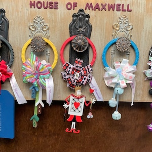 May include: Five decorative door wreaths with various themes, including a red bow, a colorful bow, a red and black playing card design, a blue and white bow, and a black and white bow with bells. The wreaths are hanging on a wooden door with the text "HOUSE OF MAXWELL" above them.