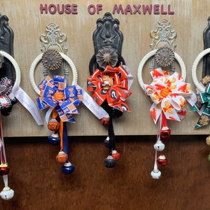 May include: Five white door knob wreaths with different colored ribbon bows and jingle bells. The ribbons are decorated with various sports team logos. The sign above reads "House of Maxwell".
