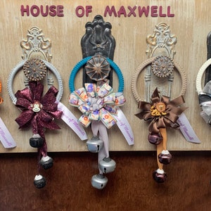May include: Five door hangers with different colored bows and bells. The door hangers are decorated with various ribbons and bows, including a tiger print, a burgundy glitter bow, a floral print, a brown bow with a gold accent, and a black and white bow. Each door hanger has a silver bell attached to a string.