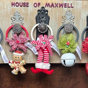 May include: A wooden sign with the text "HOUSE OF MAXWELL" features five decorative Christmas ornaments. Each ornament hangs from a decorative handle and includes a variety of festive elements like bows, bells, and a gingerbread man.