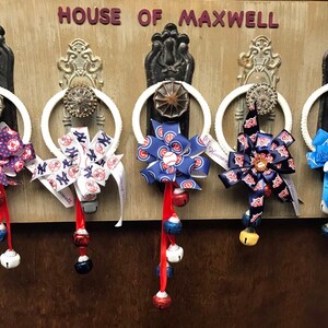 May include: Five white door knob wreaths decorated with baseball team themed pinwheels and jingle bells. The wreaths are hanging on a wooden door with the words "House of Maxwell" above them.