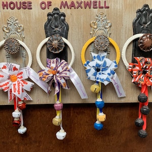 May include: Four door hangers with different sports team themed bows and jingle bells. The bows are in the colors of the Chicago Bulls, Los Angeles Lakers, a blue and white team, and a red and black team. Each hanger has a white ribbon with the text "House of Maxwell" printed on it.