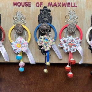 May include: A collection of decorative ring ornaments with bows and bells. Each ring is a different color, including red, yellow, blue, and white. The bows feature various patterns and ribbons with text. The ornaments hang from a wooden board with the text "HOUSE OF MAXWELL".