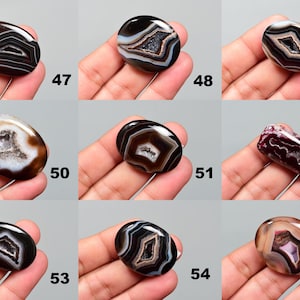 Op de afbeelding: Assortiment van 10 ovale agaat cabochons met verschillende patronen in zwart, wit en bruin. De cabochons worden getoond op een hand.