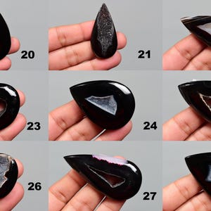 Op de afbeelding: Tien zwarte agaat cabochons met witte en grijze druzy-insluitsels. De cabochons hebben de vorm van een traan en zijn gepolijst. Ze zijn gerangschikt in drie rijen van drie, met de nummers 20 tot en met 28 gedrukt onder elk cabochon.