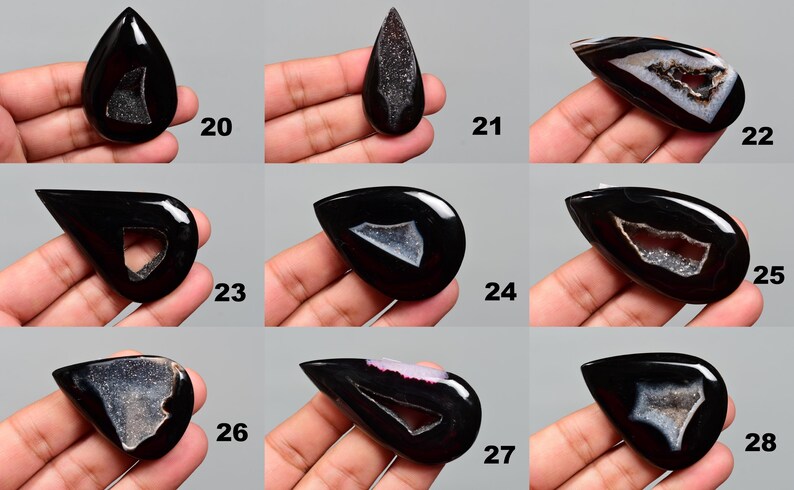 Op de afbeelding: Tien zwarte agaat cabochons met witte en grijze druzy-insluitsels. De cabochons hebben de vorm van een traan en zijn gepolijst. Ze zijn gerangschikt in drie rijen van drie, met de nummers 20 tot en met 28 gedrukt onder elk cabochon.