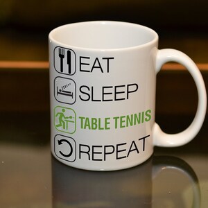 May include: White ceramic mug with black text that reads "Eat, Sleep, Table Tennis, Repeat". The text is in a list format with icons next to each item. The icon for table tennis is green.