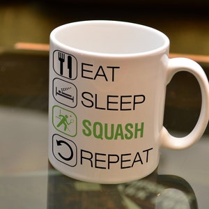May include: White ceramic mug with black text that reads "Eat Sleep Squash Repeat" with a graphic of a squash player and a squash ball in green.