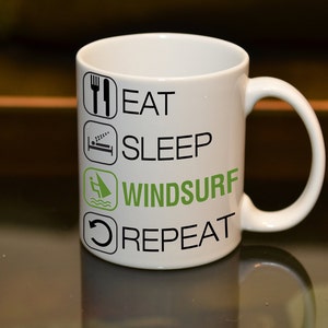May include: White ceramic mug with black text that reads "Eat, Sleep, Windsurf, Repeat". The word "Windsurf" is in green text. There is a small icon next to each word.