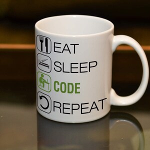 Peut inclure: Une tasse en céramique blanche avec du texte noir qui dit "Manger Dormir Coder Répéter" avec des icônes pour chaque mot. L'icône pour "Coder" est verte.
