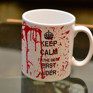 May include: White ceramic coffee mug with red blood splatter design. The mug has the text "Keep Calm I'm the Best First Aider" printed on it.