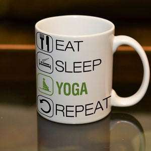 May include: White ceramic mug with the words "EAT SLEEP YOGA REPEAT" in black and green. The mug features icons of a fork and knife, a bed, a person in a yoga pose, and a circular arrow.