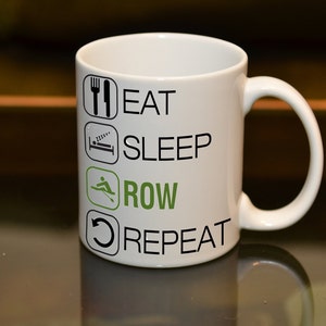 May include: White ceramic coffee mug with black text that reads "Eat, Sleep, Row, Repeat". The word "Row" is in green and features a rowing icon.