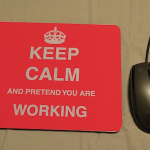 Peut inclure: Un tapis de souris rouge avec le texte "Keep Calm and Pretend You Are Working" en lettres blanches. Un graphique de couronne est au-dessus du texte. Une souris d'ordinateur noire avec le mot "Advent" dessus est sur le côté droit de l'image.
