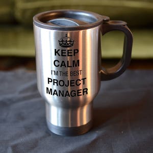May include: A silver travel mug with a black handle and lid. The mug features the text "KEEP CALM I'M THE BEST PROJECT MANAGER" in black, topped with a crown graphic.